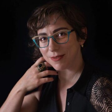 A photo of Cyndi Gilbert. Cyndi is wearing eyeglasses that are half tan and half turquoise coloured. Cyndi's hand is near her chin and there is a ring on their middle finger.
